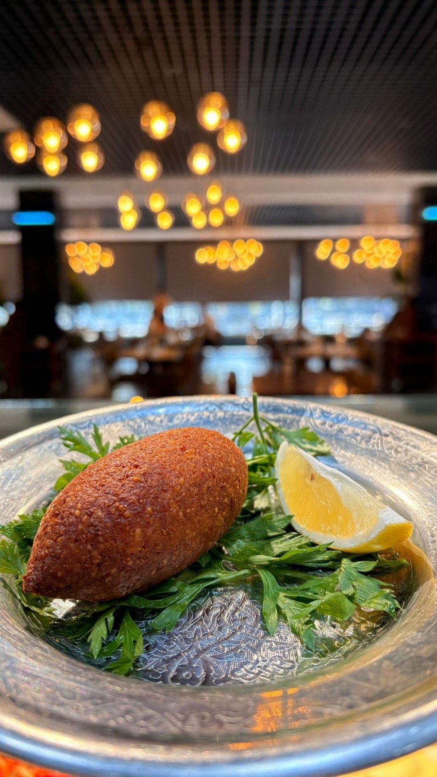 Gümüş tabakta kızarmış içli köfte, maydanoz ve limon dilimi, arkada salon