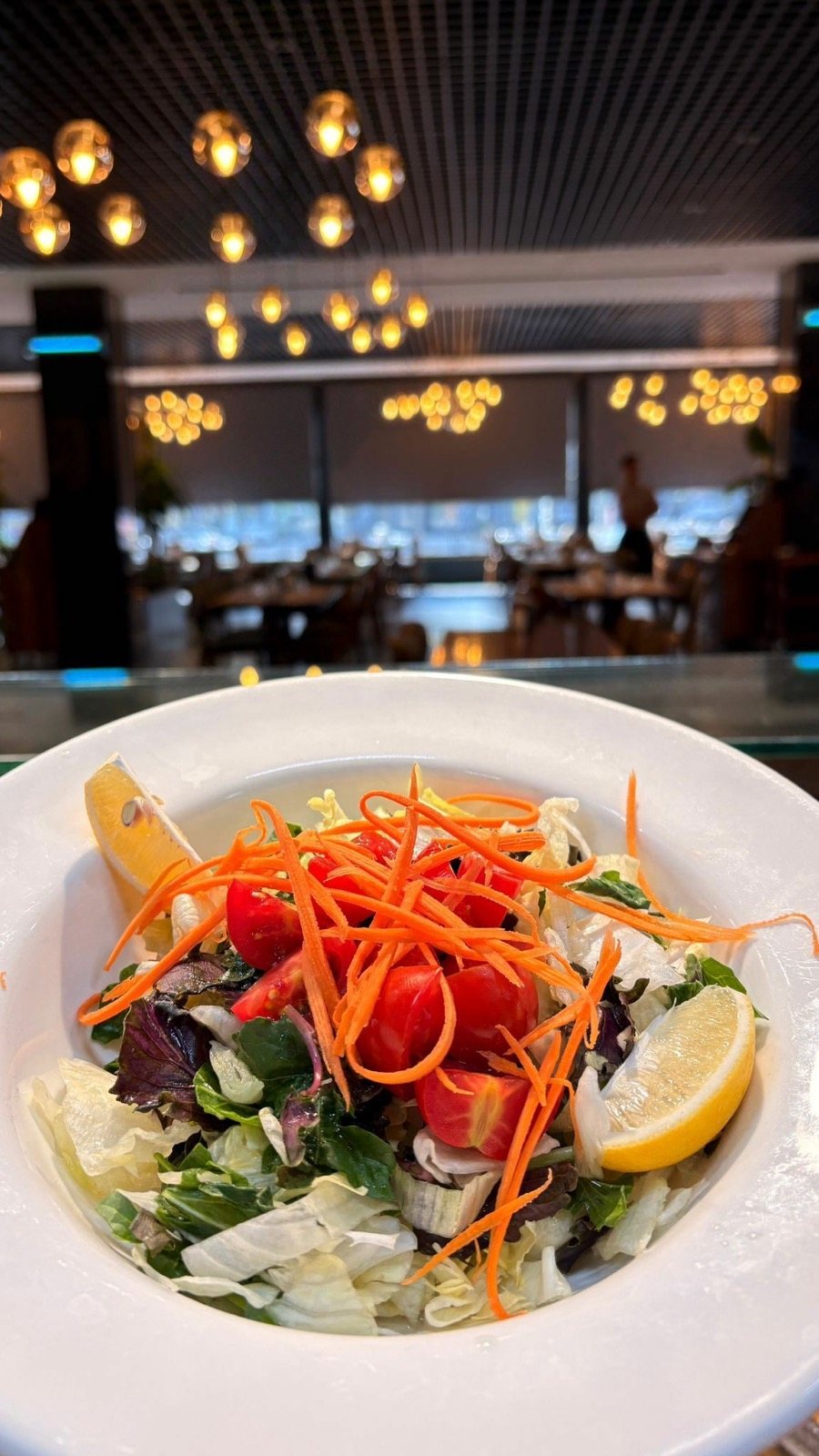 Beyaz kâsede havuçlu mevsim salatası ve limon, arkada restoran salonu