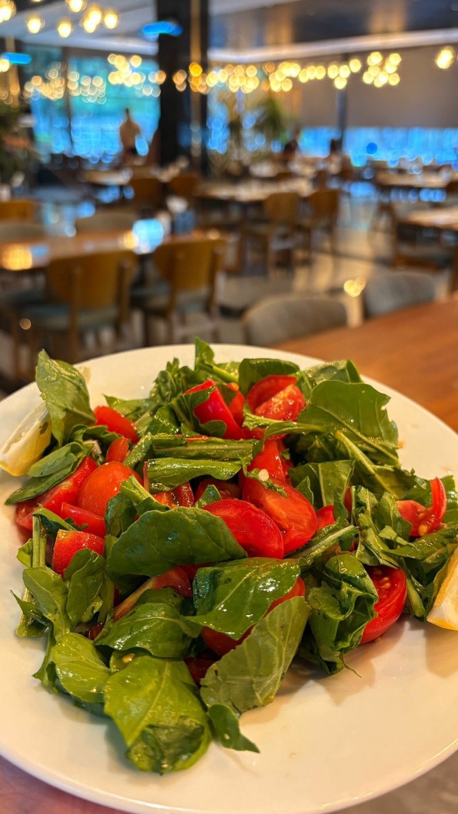Beyaz tabakta roka salatası ve domates dilimleri, arkada restoran salonu